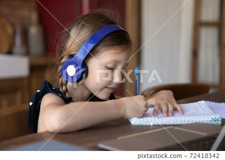 Close up little girl wearing headphones writing notes, studying online 72418342
