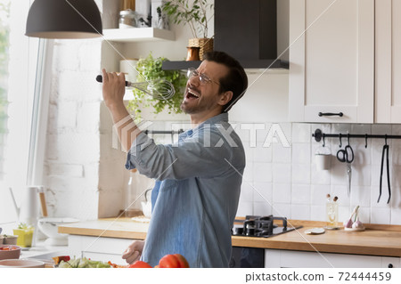 Funny man wearing glasses singing into whisk in kitchen 72444459