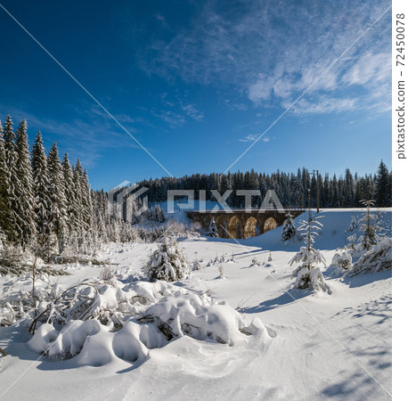 Stone viaduct (arch bridge) on railway through mountain snowy fir forest. Snow drifts on wayside and hoarfrost on trees and electric line wires. Stone viaduct (arch bridge) on railway through mountain snowy fir forest. Snow drifts on wayside and hoarfrost on trees and electric line wires. 72450078