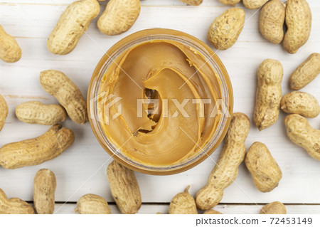 A jar of peanut butter on a white wooden table with peanuts scattered nearby. 72453149