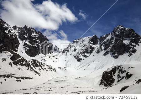 Snowy sunlit mountains with traces from avalanches and blue cloudy sky 72455112