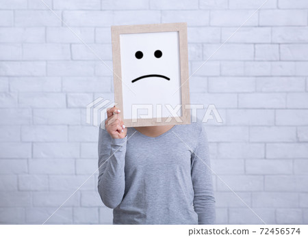 Millennial lady holding picture frame with unhappy emoticon, covering her face against white wall 72456574