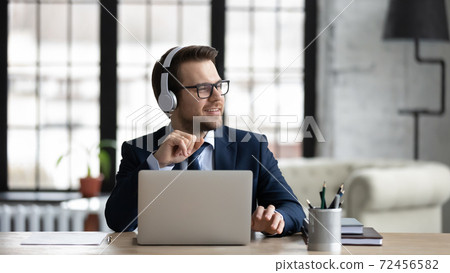 Dreamy businessman wearing headphones sitting at desk in office 72456582