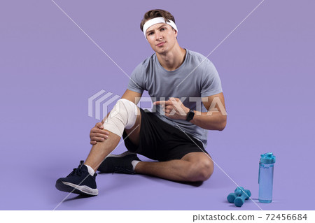 Athletic millennial man having sports injury, pointing at his sprained bandaged knee over lilac studio background Athletic millennial man having sports injury, pointing at his sprained bandaged knee over lilac studio background 72456684