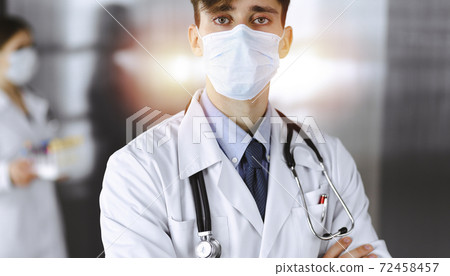 Young male doctor standing with arms crossed in clinic. Smart physician wearing medical protective mask in modern hospital. Medicine and Corona virus concept 72458457