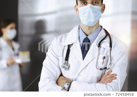 Young male doctor standing with arms crossed in clinic. Smart physician wearing medical protective mask in modern hospital. Medicine and Corona virus concept 72458458
