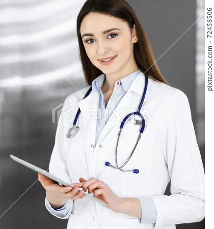 Cheerful intelligent woman-doctor is holding a tablet computer in her hands, while she is standing in a clinic. Portrait of a friendly physician woman at work. Perfect medical service in a hospital Cheerful intelligent woman-doctor is holding a tablet computer in her hands, while she is standing in a clinic. Portrait of a friendly physician woman at work. Perfect medical service in a hospital 72458506