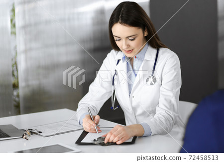 Smiling young woman-doctor is filling up medical recommendations using a clipboard. Therapist at work in a clinic office. Medicine concept 72458513