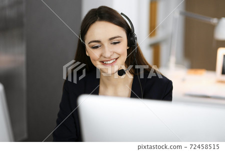 Young friendly girl in headsets is talking to a firm's client, while sitting at the desk in a modern office together with her colleague. Call center operators at work 72458515