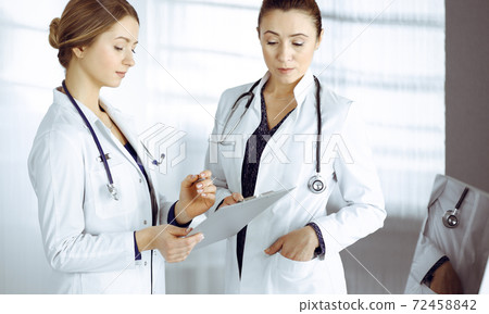 Two female physicians are discussing their patient's medical tests, while standing in a clinic office. Doctors use a clipboard at work. Teamwork in medicine Two female physicians are discussing their patient's medical tests, while standing in a clinic office. Doctors use a clipboard at work. Teamwork in medicine 72458842