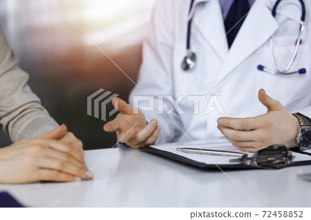 Unknown male doctor and patient woman discussing something while sittingin a darkened clinic, glare of light on the background. Close-up of hands Unknown male doctor and patient woman discussing something while sittingin a darkened clinic, glare of light on the background. Close-up of hands 72458852