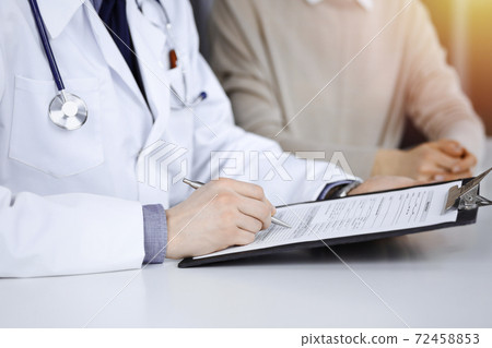Unknown male doctor and patient woman discussing something while sittingin a darkened clinic and using clipboard, glare of light on the background Unknown male doctor and patient woman discussing something while sittingin a darkened clinic and using clipboard, glare of light on the background 72458853