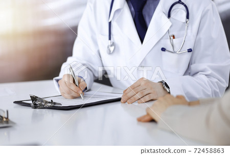 Unknown male doctor and patient woman discussing something while sittingin a darkened clinic and using clipboard, glare of light on the background Unknown male doctor and patient woman discussing something while sittingin a darkened clinic and using clipboard, glare of light on the background 72458863