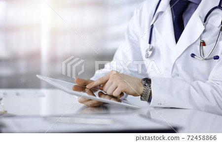 Unknown male doctor sitting and working with tablet computer iin a darkened clinic, glare of light on the background, close-up of hands Unknown male doctor sitting and working with tablet computer iin a darkened clinic, glare of light on the background, close-up of hands 72458866