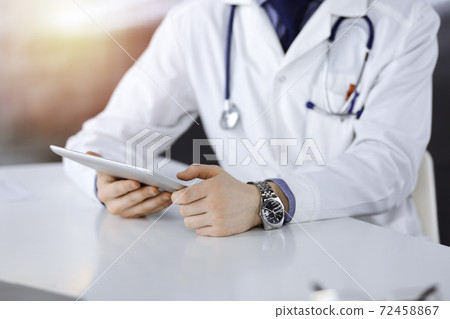 Unknown male doctor sitting and working with tablet computer iin a darkened clinic, glare of light on the background, close-up of hands Unknown male doctor sitting and working with tablet computer iin a darkened clinic, glare of light on the background, close-up of hands 72458867