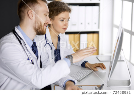 Red-bearded doctor with female colleague discussing current disease therapy while sitting at working place in clinic. Team work in medicine Red-bearded doctor with female colleague discussing current disease therapy while sitting at working place in clinic. Team work in medicine 72458981
