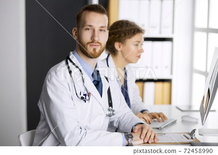 Red-bearded doctor with female colleague discussing current disease therapy while sitting at working place in clinic. Team work in medicine Red-bearded doctor with female colleague discussing current disease therapy while sitting at working place in clinic. Team work in medicine 72458990