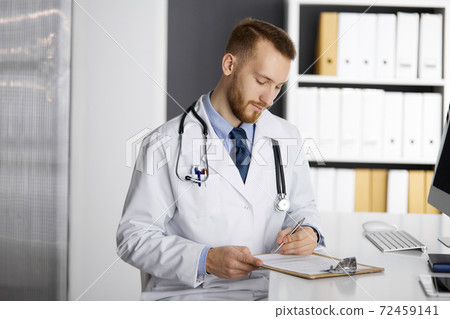 Friendly red-bearded doctor sitting and writing at clipboard in clinic. Perfect medical service in hospital. Medicine concept Friendly red-bearded doctor sitting and writing at clipboard in clinic. Perfect medical service in hospital. Medicine concept 72459141