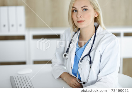 Portrate of woman-doctor at work while sitting at the desk in clinic. Blonde cheerful physician ready to help patients Portrate of woman-doctor at work while sitting at the desk in clinic. Blonde cheerful physician ready to help patients 72460031