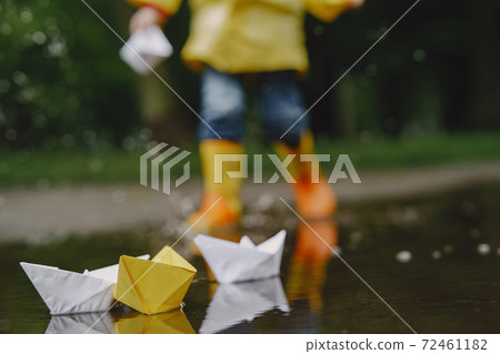 Funny kids in rain boots playing with paper ship by a puddle 72461182