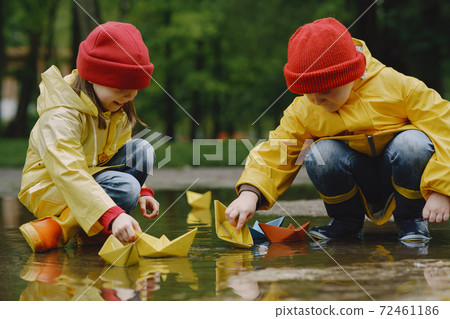 Funny kids in rain boots playing with paper ship by a puddle 72461186