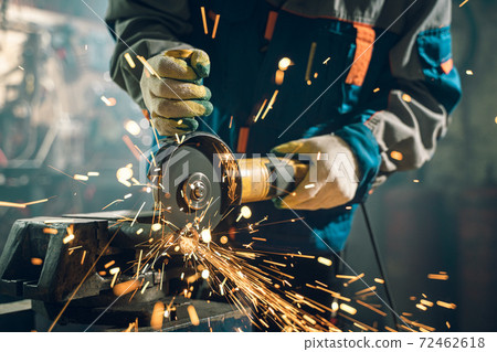 Locksmith in special clothes and goggles works in production. Metal processing with angle grinder. Sparks in metalworking 72462618