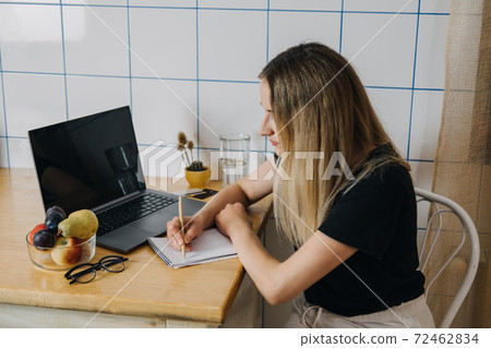 Shopping list, online Shopping, Save money. Young woman writing shopping list sitting near laptop at kitchen. Woman writing shopping list while sitting near notebook at home Shopping list, online Shopping, Save money. Young woman writing shopping list sitting near laptop at kitchen. Woman writing shopping list while sitting near notebook at home 72462834