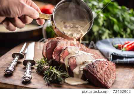 Rump steak cut to slices on a chopping board next to a knife and rosemary, with a mushroom sauce poured over it 72462903