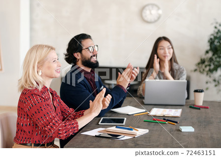 Happy intercultural professionals clapping their hands while sitting by table 72463151
