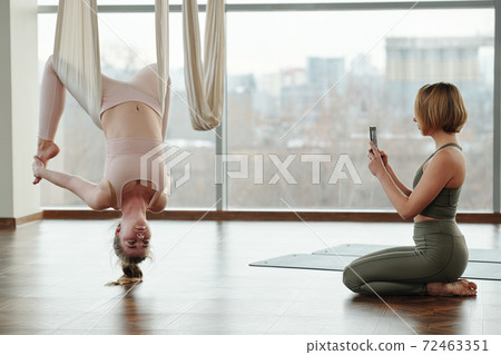 Young woman with smartphone taking photo of female in tracksuit exercising 72463351