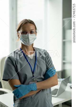 Young serious female clinician in uniform, protective eyewear, mask and gloves Young serious female clinician in uniform, protective eyewear, mask and gloves 72463715