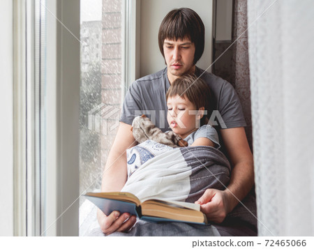 Father reads book to his son. Cozy family time on windowsill while snowfall outside. Toddler boy sits together with fluffy toy cover himself with blanket. 72465066