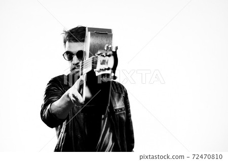 A man plays the guitar in a black leather jacket with sunglasses on a light background A man plays the guitar in a black leather jacket with sunglasses on a light background 72470810