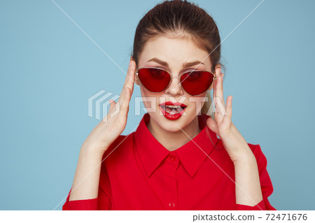 Woman in red shirt on blue background holds hands near face and sunglasses red lips makeup model Woman in red shirt on blue background holds hands near face and sunglasses red lips makeup model 72471676