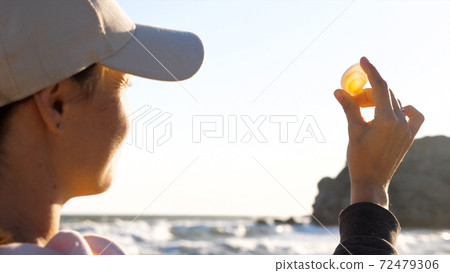 Young woman is holding seashell in hand and looking on sun through shell 72479306