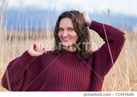 Portrait of pretty young woman in brown sweater stands in dried grass in field 72493680