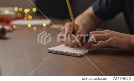 man writes christmas gift list for holidays in notepad with a pencil under warm light in the evening man writes christmas gift list for holidays in notepad with a pencil under warm light in the evening 72494690
