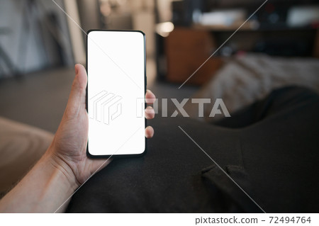 POV photo of man holding smartphone with white screen while sitting on a couch 72494764
