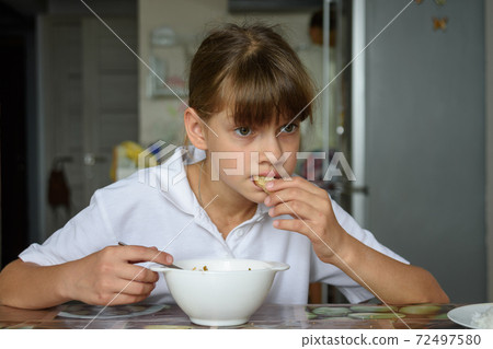 Tired hungry schoolgirl eating soup at the table in the kitchen Tired hungry schoolgirl eating soup at the table in the kitchen 72497580