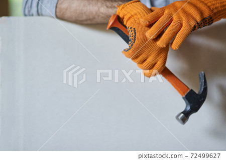Close up shot of hands of male builder wearing protective gloves holding hammer while working with drywall at construction site 72499627