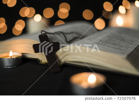 Cross placed on a open bible and candles against bokeh lights background 72507076