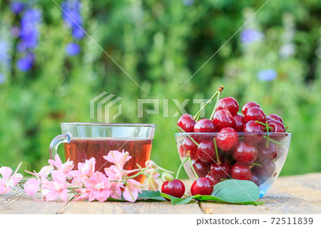 Red ripe cherry fruits with peduncles in glass bowl and cup of tea. Red ripe cherry fruits with peduncles in glass bowl and cup of tea. 72511839