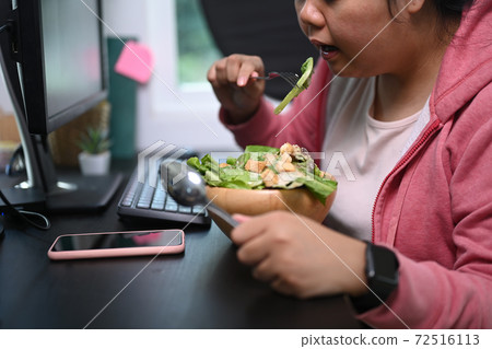 Cropped shot young overweight woman sat at table and eating healthy salad at her workspace. Cropped shot young overweight woman sat at table and eating healthy salad at her workspace. 72516113
