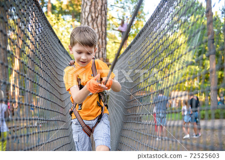 Young cute child boy in summer clothing, safety harness and helmet attached with carbine to cable moves slowly along rope way on green trees sunny bokeh background. Sport, game, leisure concept Young cute child boy in summer clothing, safety harness and helmet attached with carbine to cable moves slowly along rope way on green trees sunny bokeh background. Sport, game, leisure concept 72525603