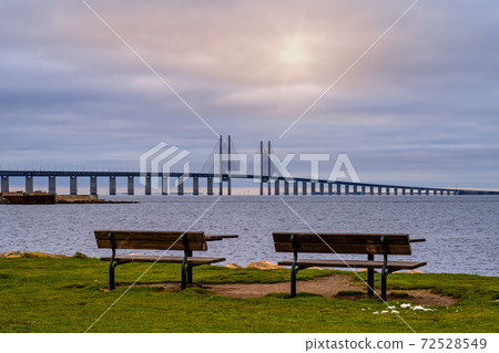 Two benches in front of a beautiful ocean view with a bridge. Blue ocean and a sunset sky in the background. Picture from Malmo, Sweden Two benches in front of a beautiful ocean view with a bridge. Blue ocean and a sunset sky in the background. Picture from Malmo, Sweden 72528549