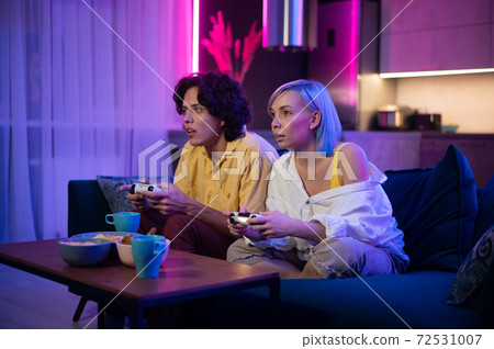 Happy boyfriend and girlfriend playing video games and holding joysticks at home. Millennial couple enjoying game while sitting on sofa and spending free time. Concept of relationship 72531007