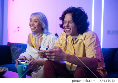 Happy boyfriend and girlfriend playing video games and holding joysticks at home. Millennial couple enjoying game while sitting on sofa and spending free time. Concept of relationship 72531018