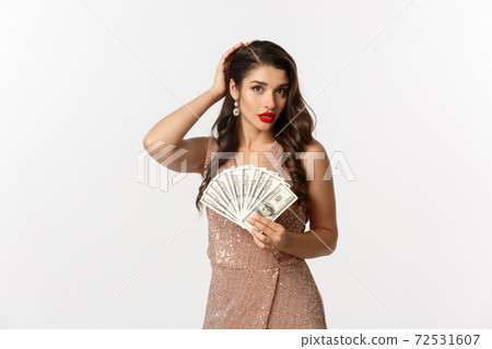 Shopping concept. Beautiful and successful woman in elegant dress, fixing hairstyle and holding money, looking perfect, standing over white background 72531607