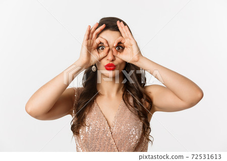 Concept of New Year celebration and winter holidays. Close-up of beautiful brunette woman in dress, red lips, making hand binoculars and staring at camera, white background 72531613
