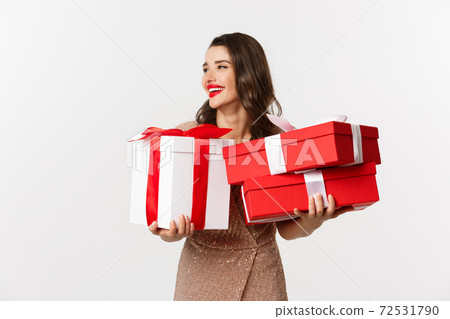 Holidays, celebration concept. Beautiful caucasian woman in elegant dress holding Christmas presents and smiling happy, standing over white background 72531790